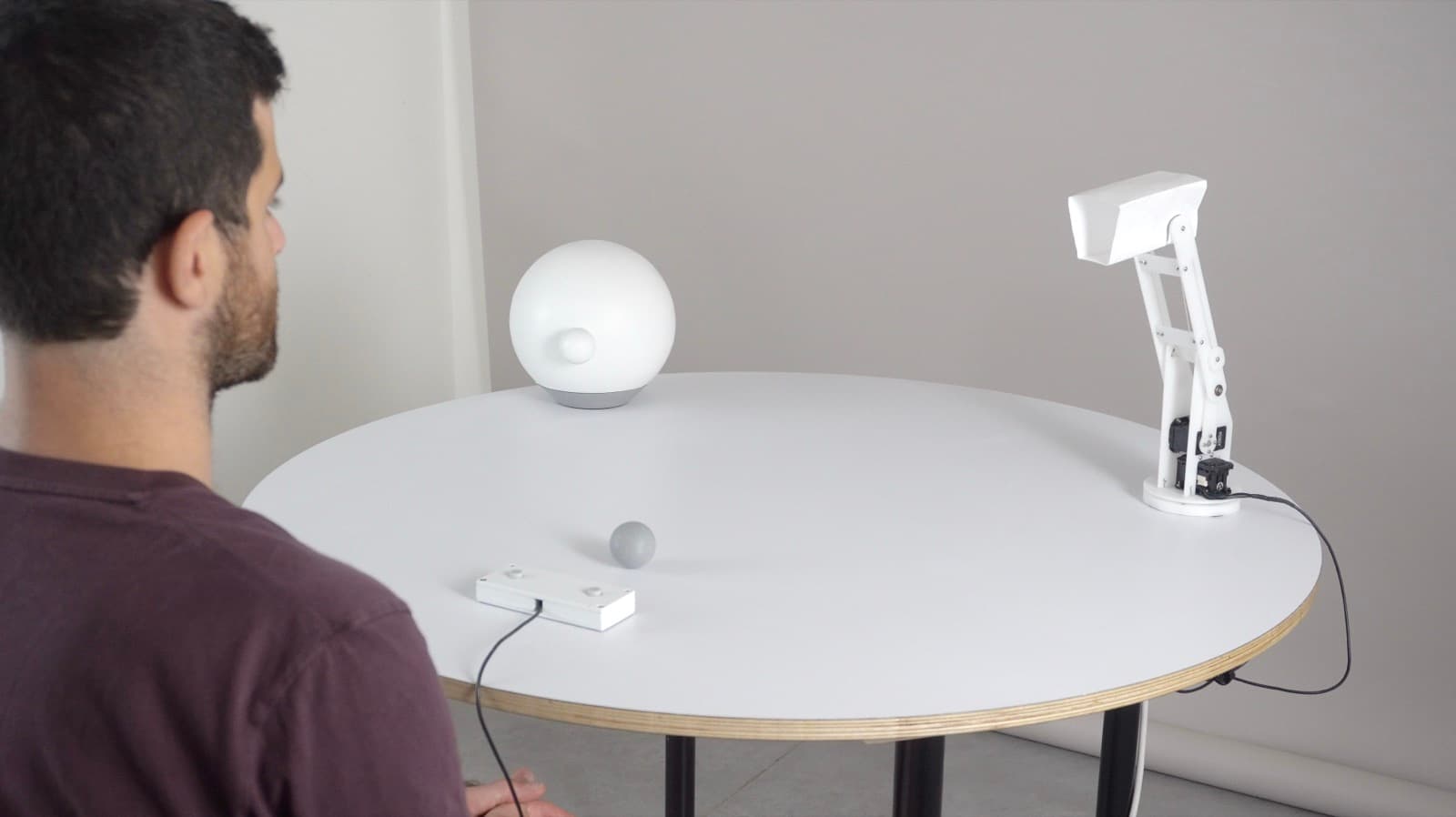 A study participant seated at a round table facing two non-humanoid robots, a sphere and a lamp-like arm, during the ball-tossing game.